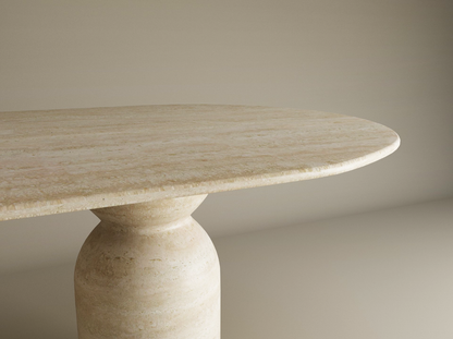 A close-up of the TRAVERTABLE Oval Travertine Dining Table, featuring an organic stone tabletop and sculptural round base, set against a plain, light-colored background. Customizable design.
