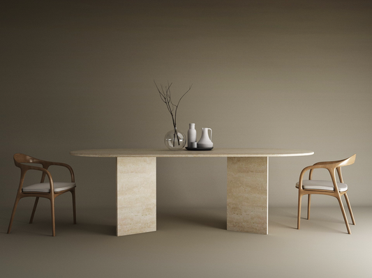 A minimalist scene features the TRAVERTABLE Oval Travertine Dining Table with Split-V Pedestal Base, paired with cushioned wooden chairs, neutral decor like a branch in a glass vase and ceramic pitchers, all enhanced by a beige backdrop.