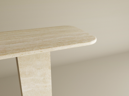 A close-up of the TRAVERTABLE Rectangular Travertine Console Table shows its light beige travertine stone top with rounded edge and rectangular pedestal base, set against a plain matching background.