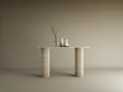 A TRAVERTABLE Ovale Travertine Console Table with Vertical Groove Base stands against a beige wall, topped with three vases—clear, white, off-white—and bare branches, creating a sculptural centerpiece in this minimal scene.
