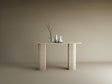 The TRAVERTABLE Ovale Travertine Console Table features a solid, fluted round base and natural stone texture. Against a beige backdrop, three minimalist vases—one with bare branches—adorn its surface in a modern, understated style.