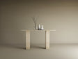 A minimalist scene with the TRAVERTABLE Rectangular Travertine Console Table, featuring a travertine stone base and top, styled with a clear vase of branches, a white pitcher, and cup against a plain beige background.
