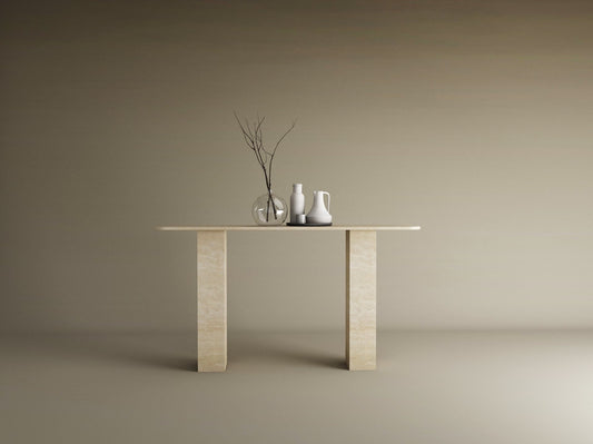 A minimalist scene with the TRAVERTABLE Rectangular Travertine Console Table, featuring a travertine stone base and top, styled with a clear vase of branches, a white pitcher, and cup against a plain beige background.
