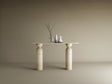 The Ovale Travertine Console Table by TRAVERTABLE, featuring a sculptural round base in organic travertine stone, displays a thin branch in a glass vase, a white pitcher, and jug against a beige backdrop—an understated statement piece.