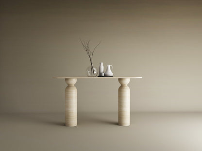 The Ovale Travertine Console Table by TRAVERTABLE, featuring a sculptural round base in organic travertine stone, displays a thin branch in a glass vase, a white pitcher, and jug against a beige backdrop—an understated statement piece.
