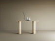 The TRAVERTABLE Ovale Travertine Console Table features a pure travertine stone base with two smooth cylindrical legs, displaying three vases—one clear with bare branches and two opaque white—set against a plain beige background.