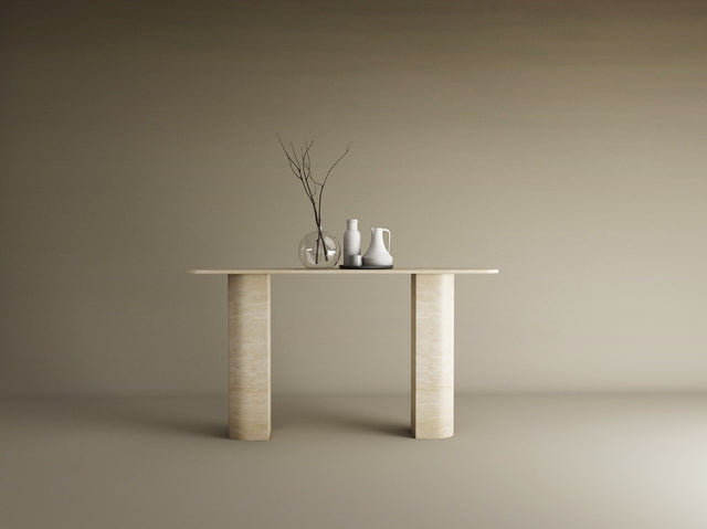 The TRAVERTABLE Rectangular Travertine Console Table with Sloped Pedestal Base in natural stone displays a glass vase with bare branches and two ceramic pitchers, set against a plain beige background.