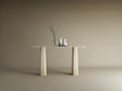 The TRAVERTABLE Rectangular Travertine Console Table with a solid, tapered round base displays a glass vase and ceramic vessels in minimalist beige, set against a plain background. This customizable piece offers contemporary elegance.