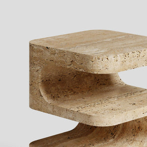 A close-up of the Serenity Block Travertine Table by Travertable reveals its C-shaped design with smooth, rounded edges and a natural stone texture against a plain light gray background. This elegant table melds functionality with timeless style.