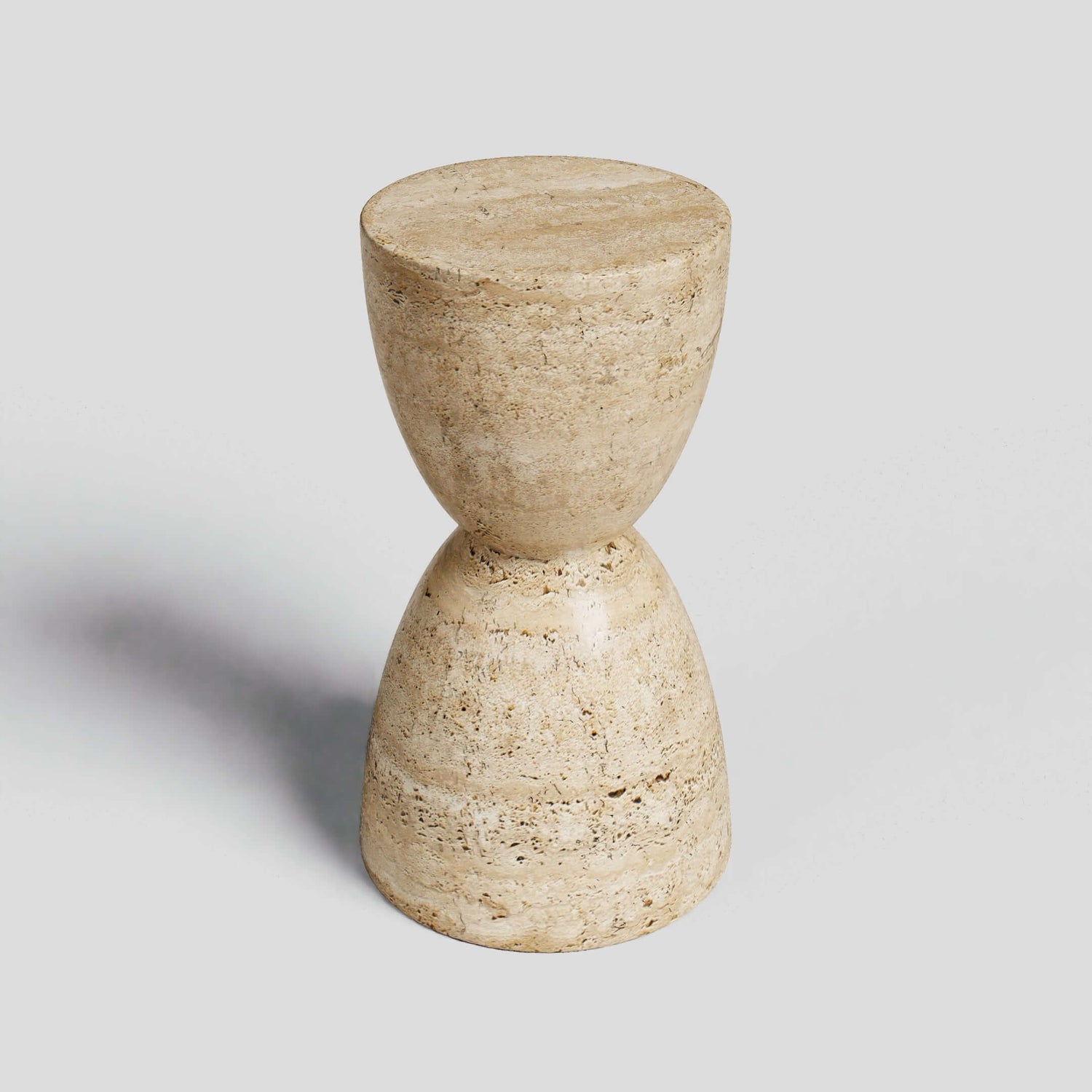 The Eda Beige Travertine Sphere Table by Travertable is a stone, hourglass-shaped stool with a rough texture, embodying minimalist design. Its symmetrical halves unite at the center, forming an abstract sculpture. Positioned against a plain gray backdrop, it casts a delicate shadow.