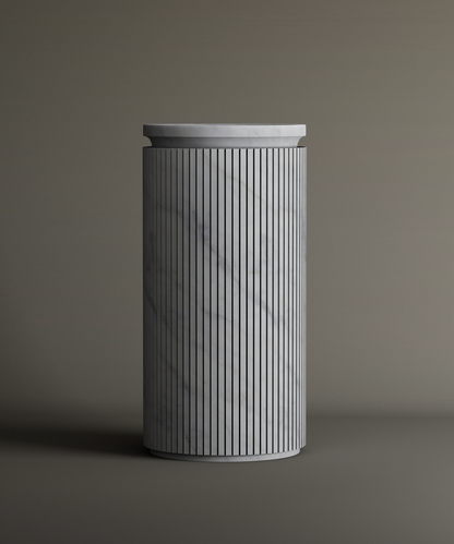 The TRAVERTABLE Carrara Round Marble Pedestal Vanity Sink features a tall, cylindrical form with vertical grooves and smooth Carrara marble surface, highlighted by subtle gray veining against a neutral background.