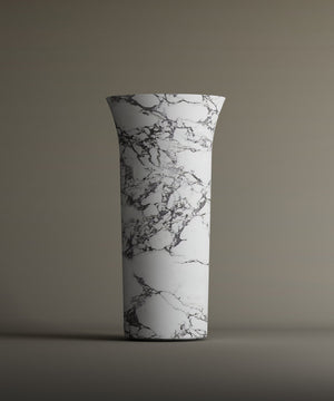 The TRAVERTABLE Calacatta Marble Cylinder Pedestal Vanity Sink features a tall, cylindrical white form with a flared rim and black-gray marble pattern, creating an elegant architectural statement against a neutral backdrop.