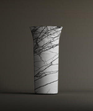 The TRAVERTABLE Lilac Marble Cylinder Pedestal Vanity Sink showcases striking black veining on white marble with a flared rim, set against a plain brownish-gray background.