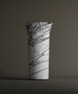 The TRAVERTABLE Lilac Marble Cylinder Pedestal Vanity Sink showcases striking black veining on white marble with a flared rim, set against a plain brownish-gray background.