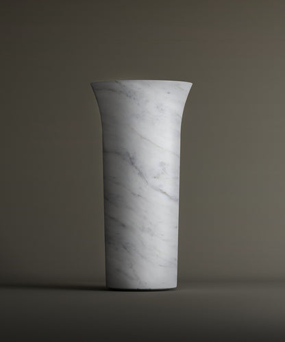 The TRAVERTABLE Carrara Marble Cylinder Pedestal Vanity Sink, with a tall cylindrical shape, white marble finish, and subtle gray veining, stands upright against a plain background. It features a slightly flared rim for added elegance.