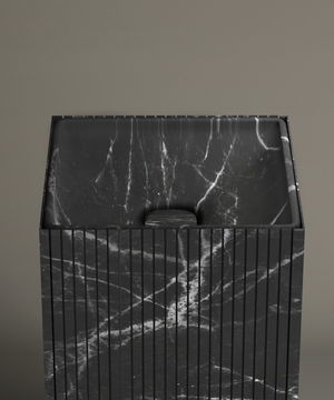 The TRAVERTABLE Nero Marquina Square Fluted Marble Pedestal Vanity Sink, featuring black marble with white veining and vertical grooves, stands against a taupe bathroom wall. Its drain cover matches the sink’s distinctive marble pattern.