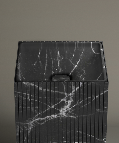 The TRAVERTABLE Nero Marquina Square Fluted Marble Pedestal Vanity Sink, featuring black marble with white veining and vertical grooves, stands against a taupe bathroom wall. Its drain cover matches the sink’s distinctive marble pattern.