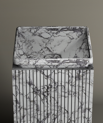 The TRAVERTABLE Calacatta Square Marble Pedestal Vanity Sink, with gray and black veining and vertical grooves, creates a bold marble centerpiece against a plain gray background.