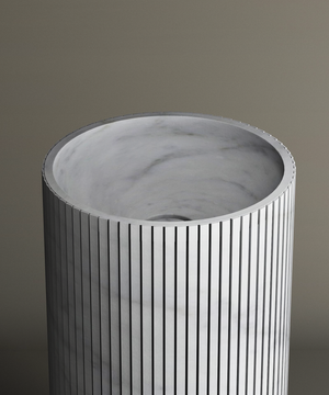 The TRAVERTABLE Carrara Round Fluted Marble Pedestal Vanity Sink features a cylindrical, fluted design with a marble texture and an open top revealing a smooth light gray interior, all set against a neutral background.