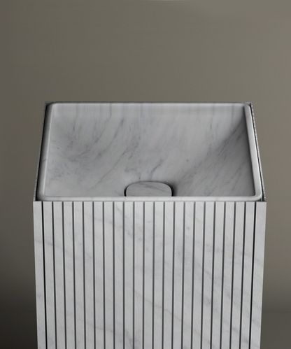 The TRAVERTABLE Carrara Square Fluted Marble Pedestal Vanity Sink features vertical grooves, a smooth angled basin, and a minimalist design—ideal for contemporary bathrooms with neutral backgrounds.