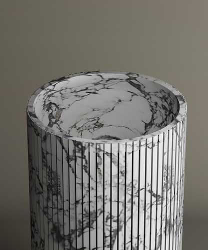 The TRAVERTABLE Calacatta Round Fluted Marble Pedestal Vanity Sink features gray veining and a fluted cylindrical exterior, standing out against a taupe backdrop with its consistent modern marble design.