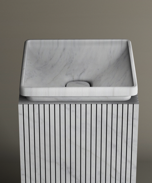 The TRAVERTABLE Carrara Square Marble Pedestal Vanity Sink features a smooth rectangular basin atop a fluted, square pedestal, serving as an elegant centerpiece against a taupe backdrop—ideal for sophisticated interiors.