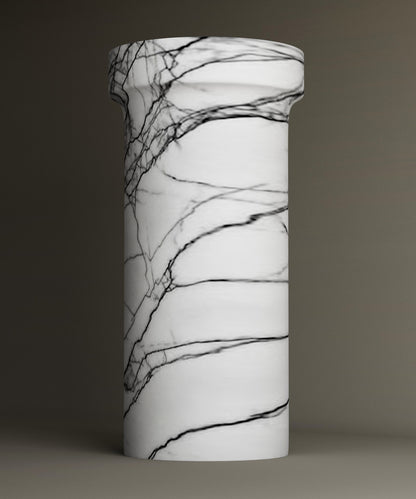 The TRAVERTABLE Lilac Marble Pedestal Vanity Sink features a freestanding cylindrical design with a white base and striking dark veining, shown against a plain, neutral background.