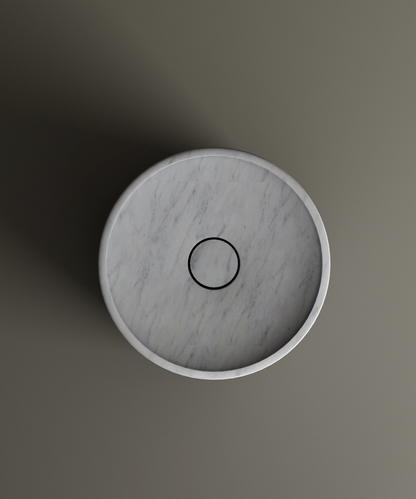 A top-down view of the TRAVERTABLE Carrara Round Marble Pedestal Vanity Sink with a black drain, set on a plain gray background. Light casts a shadow to the left, highlighting Italian-inspired elegance.