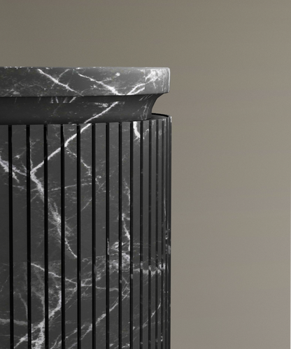 A close-up of the TRAVERTABLE Nero Marquina Round Marble Pedestal Vanity Sink, featuring vertical grooves and white veining, elevated against a beige background—an ideal centerpiece for modern bathroom design.