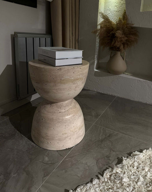 A small Eda Beige Travertine Sphere Table by Travertable, with books on top, sits next to a textured vase of dried pampas grass. The tiled floor and visible radiator in the dim room enhance the charm of minimalist furniture.