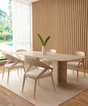 A modern dining room features the TRAVERTABLE Rectangular Travertine Dining Table with Split-V Pedestal Base, four matching chairs, a white vase with green stems centerpiece, wood slat wall panels, large window, and light rug on wood flooring.
